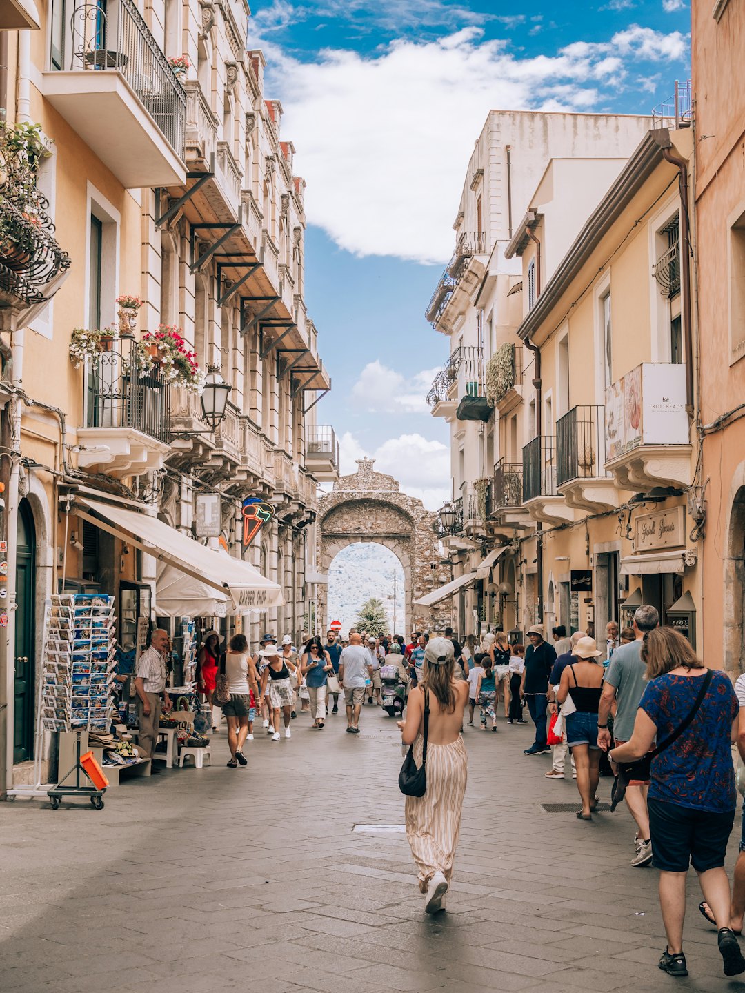 Pedestrians walk along a historic street lined with flower-filled balconies.